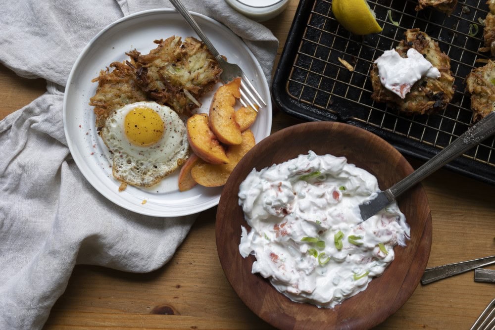 white plate with crispy potato latkes alongside over easy egg and peach slices - wood bowl with sour cream lox sauce - more latkes on cookie sheet in back