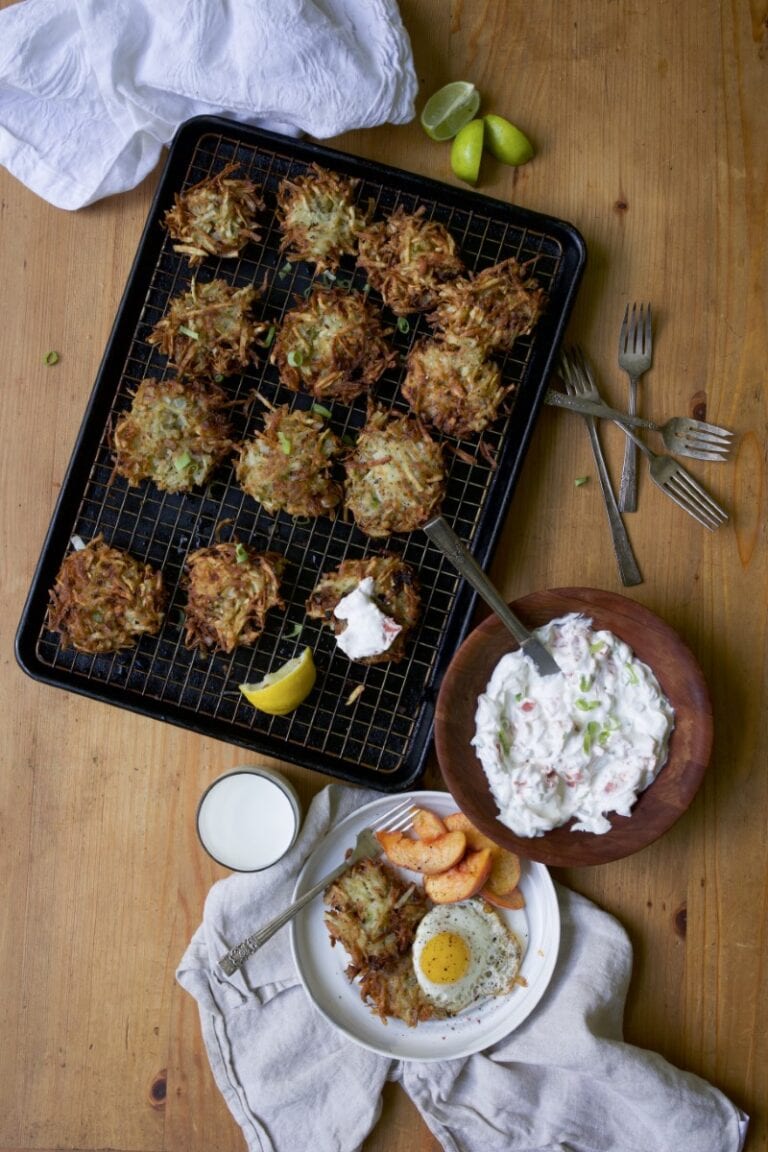 potato latkes on wire rack on baking sheet on rustic wood table - bowl with creamy lox sauce and plate with serving of latkes, fried egg, and peach slices - silverware and rumpled white dish cloths