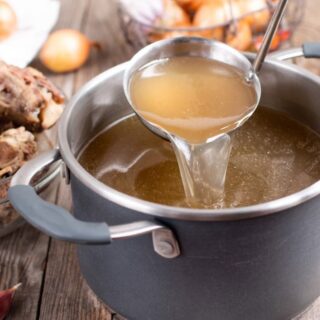 stock pot with homemade beef or venison stock, ladle lifted with spoonful, onions and bones in background