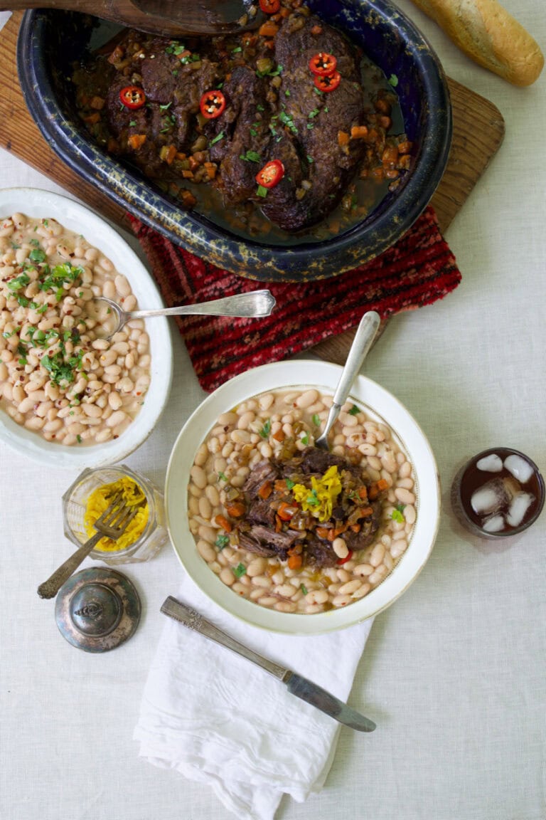 Pot Roast served in white bowl on a bed of northern beans with serving dishes of pot roast and beans in background