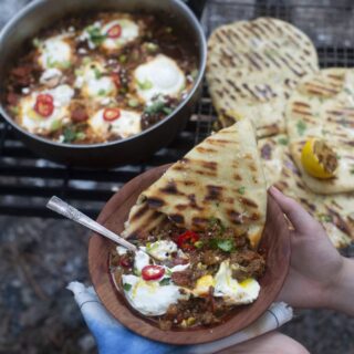 shakshuka recipe - cooked eggs with tomato sauce, served with sliced peppers, side of yogurt, and garlic naan, held up on plate with skillet in background