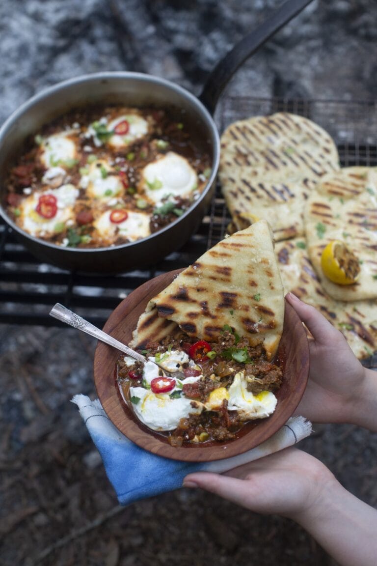 shakshuka recipe - cooked eggs with tomato sauce, served with sliced peppers, side of yogurt, and garlic naan, held up on plate with skillet in background