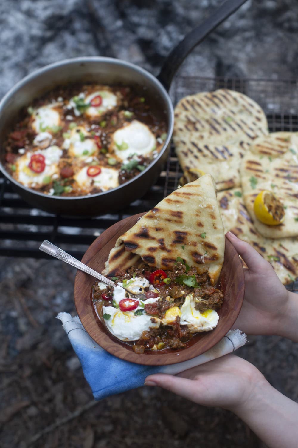 shakshuka recipe - cooked eggs with tomato sauce, served with sliced peppers, side of yogurt, and garlic naan, held up on plate with skillet in background