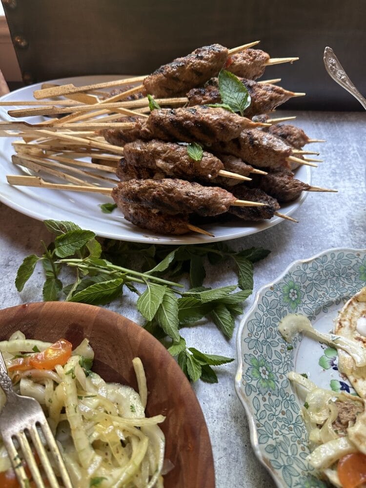 adana kebabs - venison and beef kofta kebabs on wooden skewers on a white plate behind sumac onion salad next to china plate with serving of both