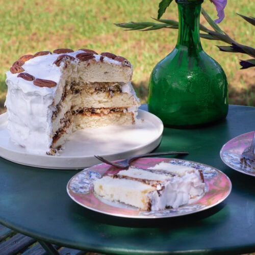 Alabama Lane Cake, a rich 3-layer cake with white icing, set on outdoor table with a wedge cut out and a serving on pink plate