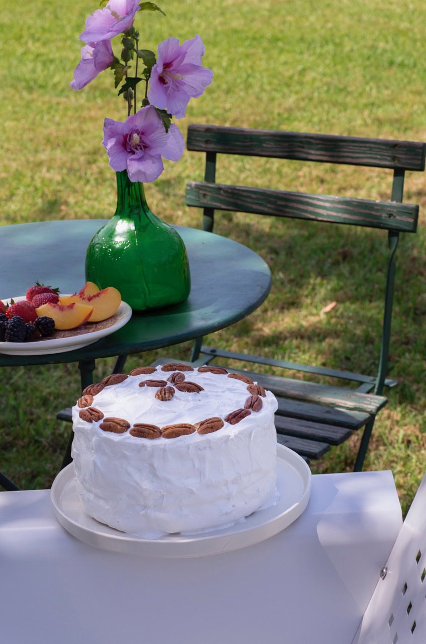 uncut creamy white Lane Cake iced and garnished with pecans, resting on table outdoors with vase of flowers on table in background