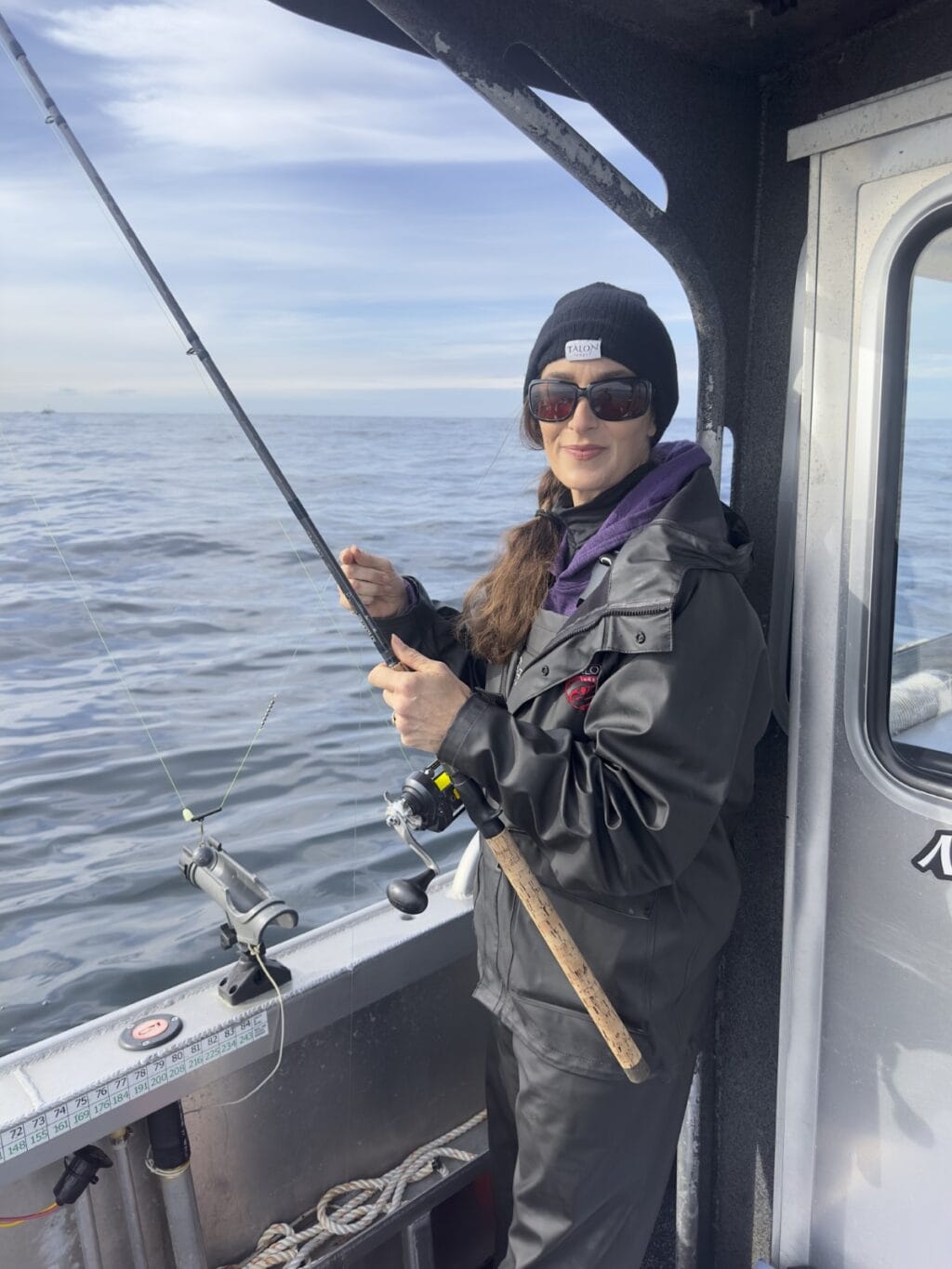 Stacy Lyn in jacket, hat, sunglasses with fishing rod on boat