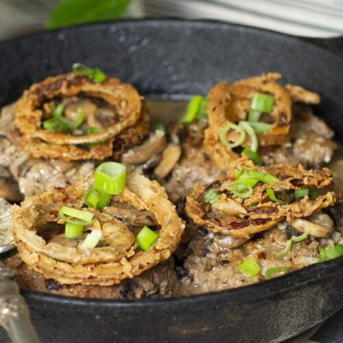 closeup of fried onion rings on top of hamburger steaks in a cast-iron skillet