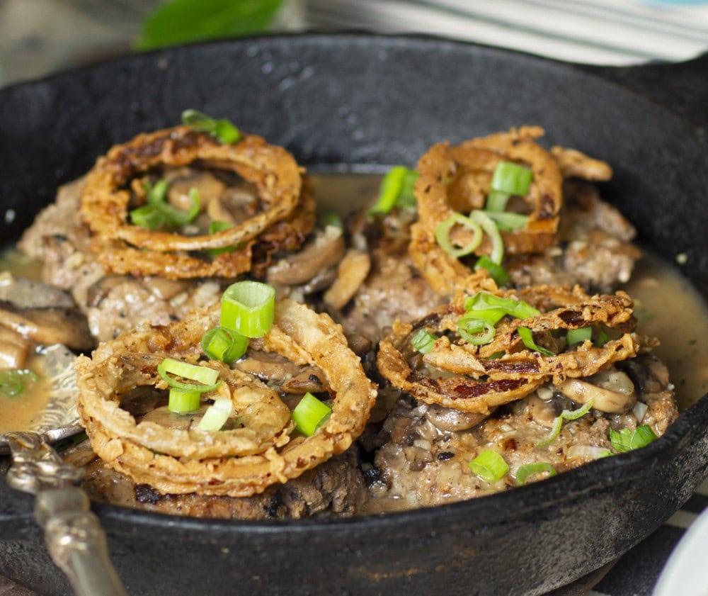 closeup of fried onion rings with green onion garnish atop hamburger steaks in cast-iron skillet