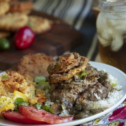 plated dinner on floral tablecloth: hamburger steak with homemade onion rings, served over rice, with mason jar of iced tea and cutting board with more onion rings in background