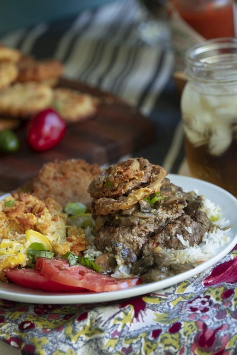plated dinner on floral tablecloth: hamburger steak with homemade onion rings, served over rice, with mason jar of iced tea and cutting board with more onion rings in background