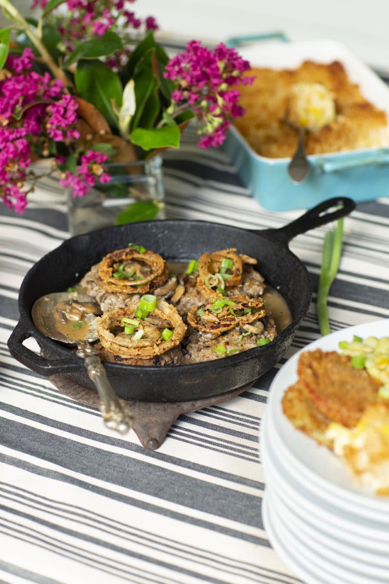 cast iron skillet with hamburger steaks in mushroom gravy, topped with crispy fried onions - pink wildflower arrangement in clear square vase in background alongside blue casserole dish