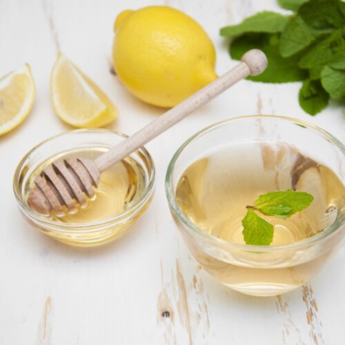 cup of lemon tea in glass cup alongside container of honey with drizzler, lemons and mint in background