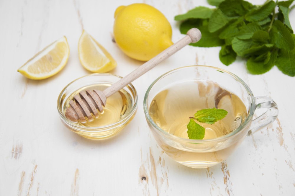 cup of lemon tea in glass cup alongside container of honey with drizzler, lemons and mint in background