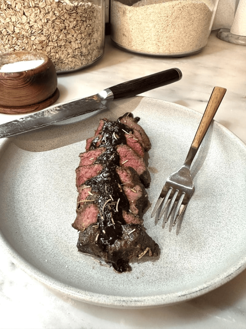 Venison medallions with mushroom wine sauce plated on white stoneware with knife and fork resting on plate, grain canisters in background