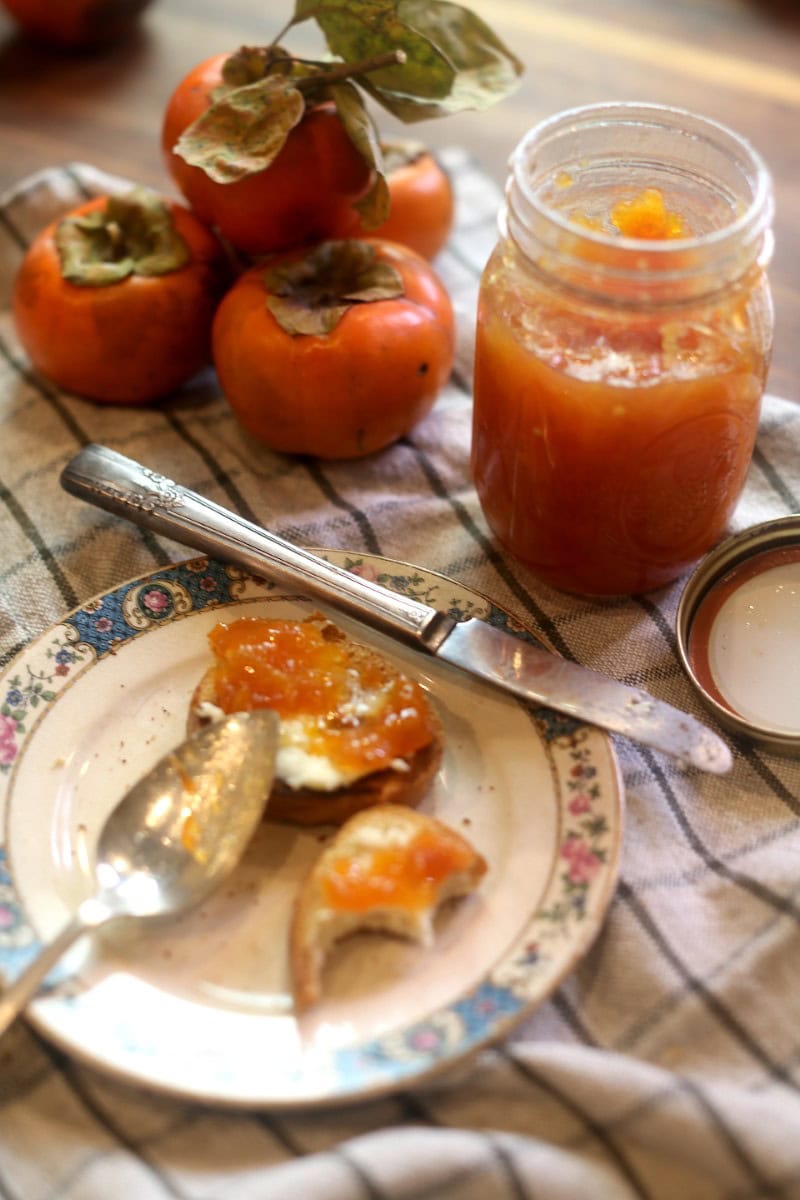 Persimmon jam spread on bagel with jar of homemade jam in background alongside a few fresh persimmons on kitchen towel