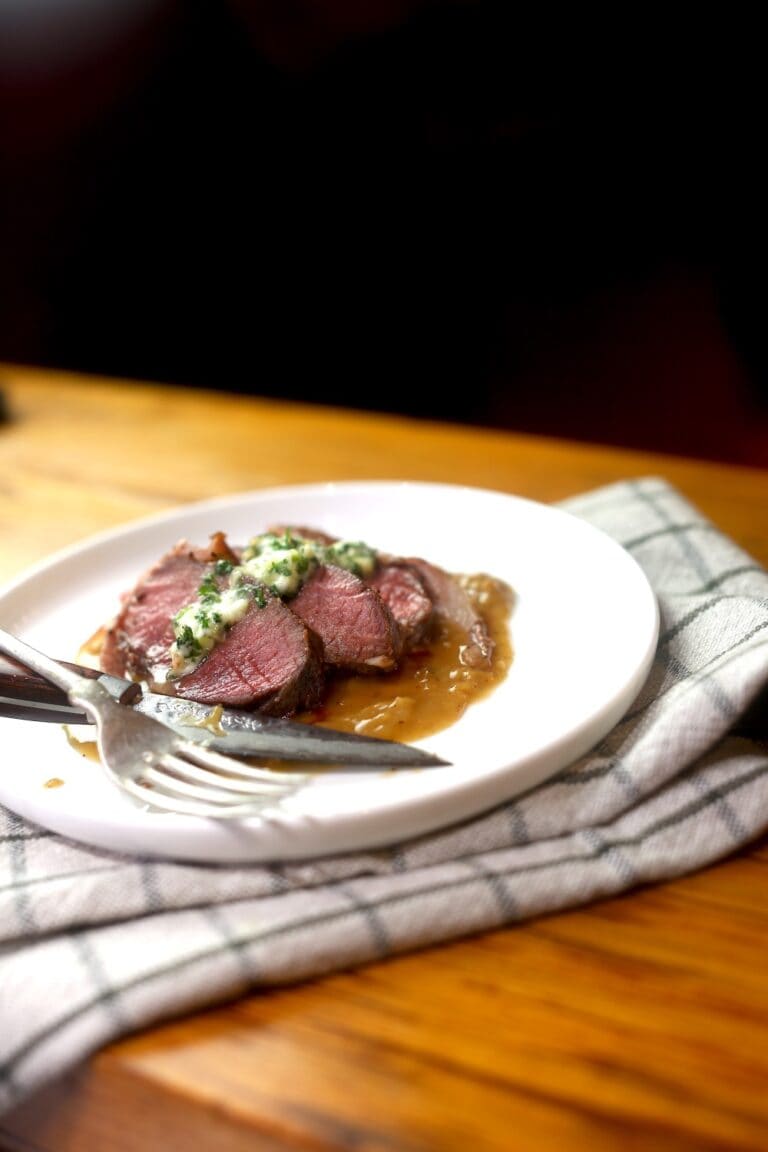 plated venison medallions, served with herb butter and gravy on white plate with napkin underneath, steak knife and fork resting on plate