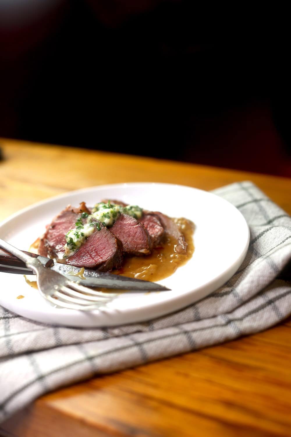 plated venison medallions, served with herb butter and gravy on white plate with napkin underneath, steak knife and fork resting on plate
