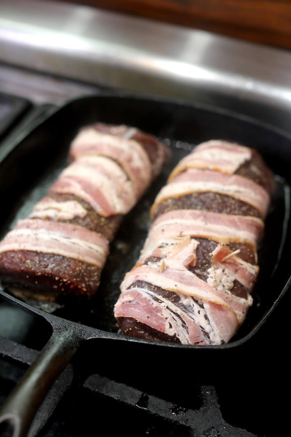 venison backstrap halves wrapped in slices of bacon to cook in cast-iron skillet