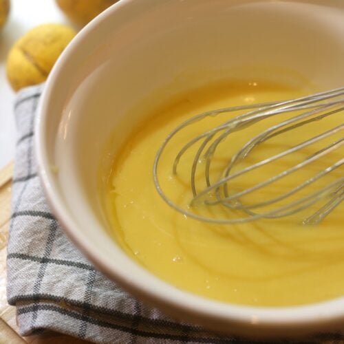 bright yellow lemon curd being whisked in large bowl with whole lemons in background