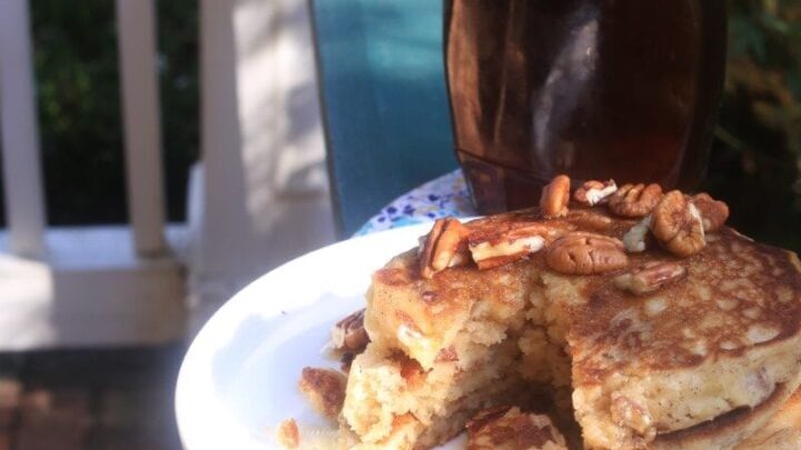 stack of fluffy pecan pancakes on white plate with floral napkin, resting on porch railing with bottle of syrup