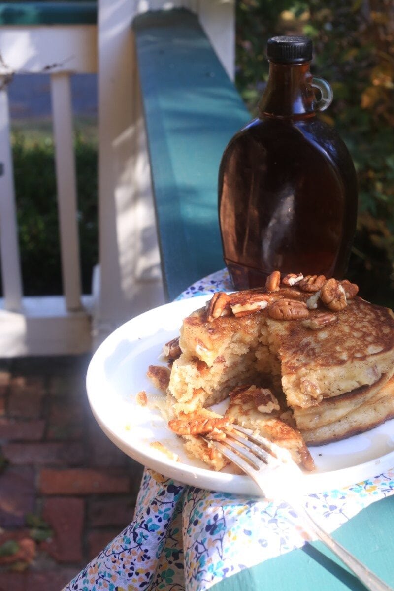 stack of fluffy pecan pancakes on white plate with floral napkin, resting on porch railing with bottle of syrup