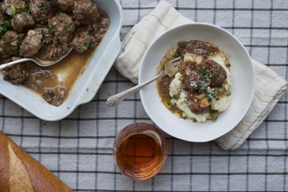 meatballs with gravy served in white bowl alongside pan of meatballs and French bread, glass of tea
