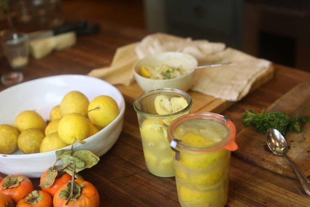 on wooden counter, jars of preserved lemons in foreground with white bowl of fresh whole lemons, bowl of pasta, fresh parsley on chopping board