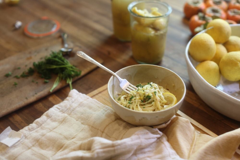 bowl of pasta on counter with jars of preserved lemons, bowl of fresh lemons, chopped parsley in background