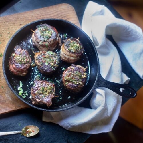 venison tenderloin in skillet resting on cutting board with white towel, dish of herb butter