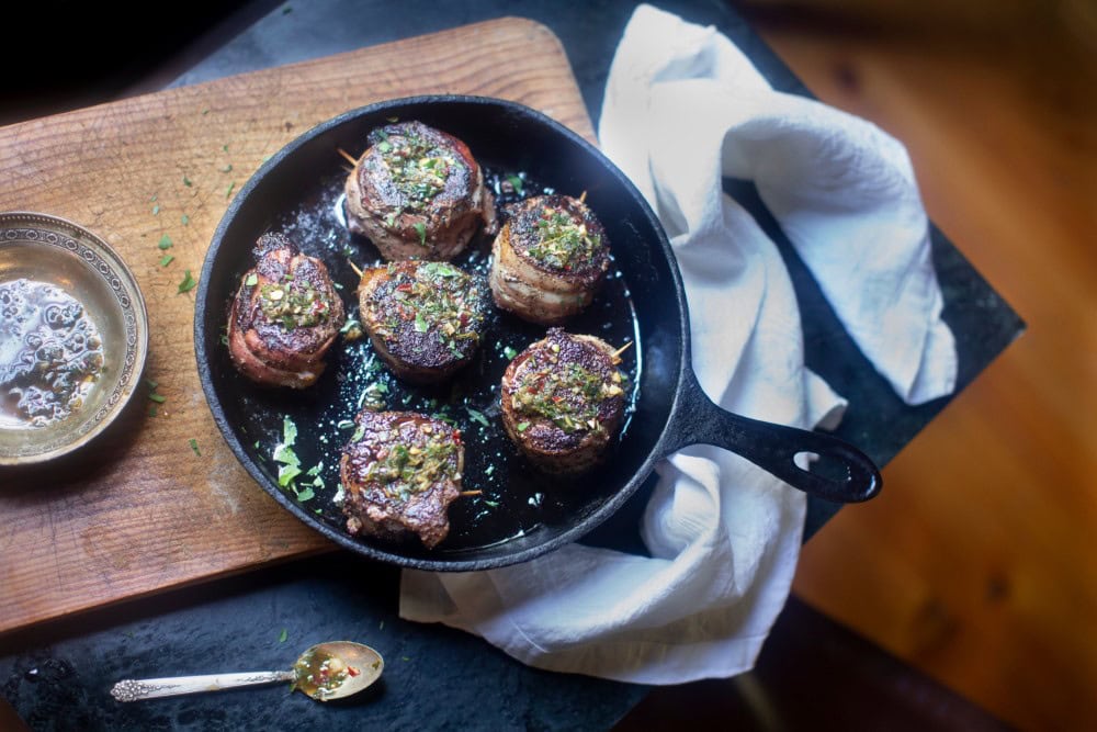 venison tenderloin in skillet resting on cutting board with white towel, dish of herb butter