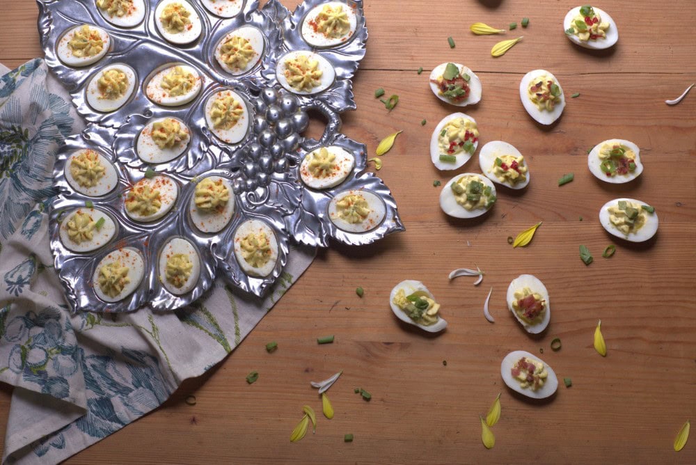 deviled eggs with a variety of toppings in a silver leaf-shaped deviled egg platter on floral napkin and spread across wood countertop