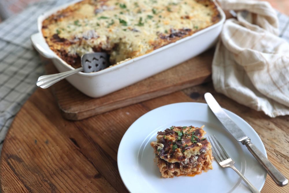 square serving of Greek lasagna - pastitsio - showing the tubular shape of the pasta. Baking dish in background shows creamy sauce.
