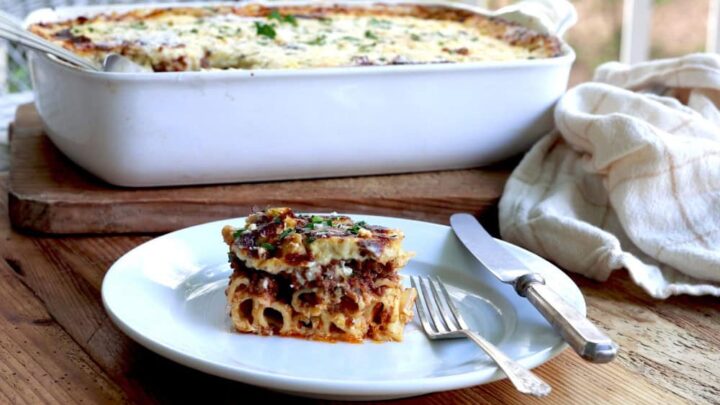 Greek lasagna served up on white plate on wooden porch table with casserole dish in background
