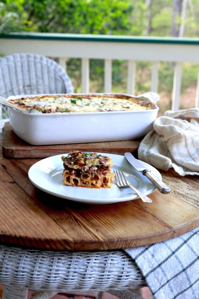 Greek lasagna served up on white plate on wooden porch table with casserole dish in background