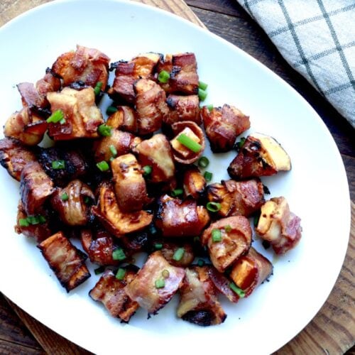 baked wrapped sweet potato bites on white oval platter sitting on rustic wooden cutting board next to checked white dish towel