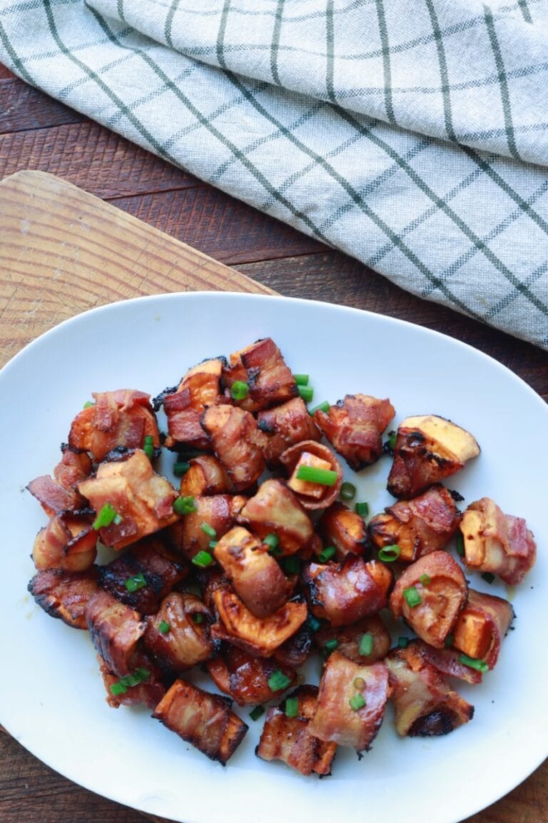 Bacon wrapped sweet potato bites on a white plate, garnished with chopped green onions.