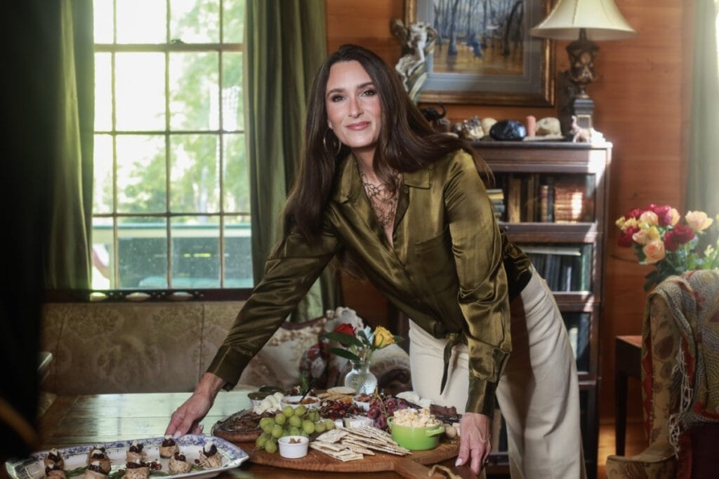 Stacy Lyn placing wild turkey roll ups on dining room table alongside a charcuterie board loaded with cheeses, crackers, and fruit